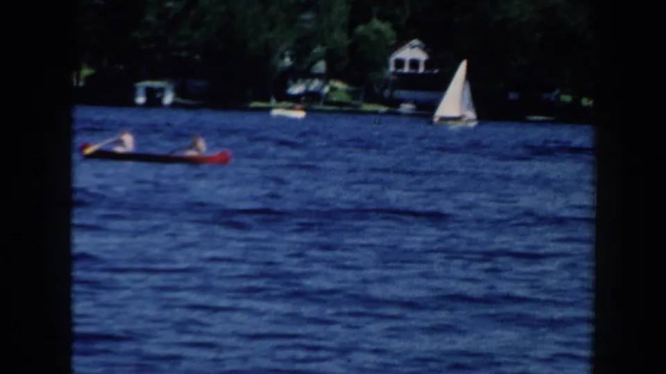 1960 Brighton, Michigan frame showing the red canoe partly near the frame edge while a white sailboat and wooded lakeside houses remain visible.