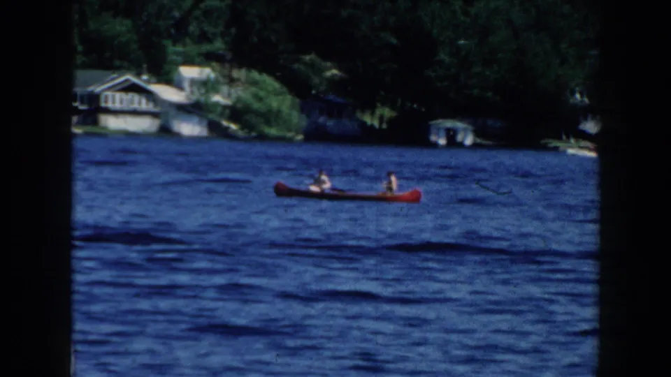 1960 Brighton, Michigan home-movie frame of two people in a red canoe on blue lake water, with wooded shoreline houses in the background.