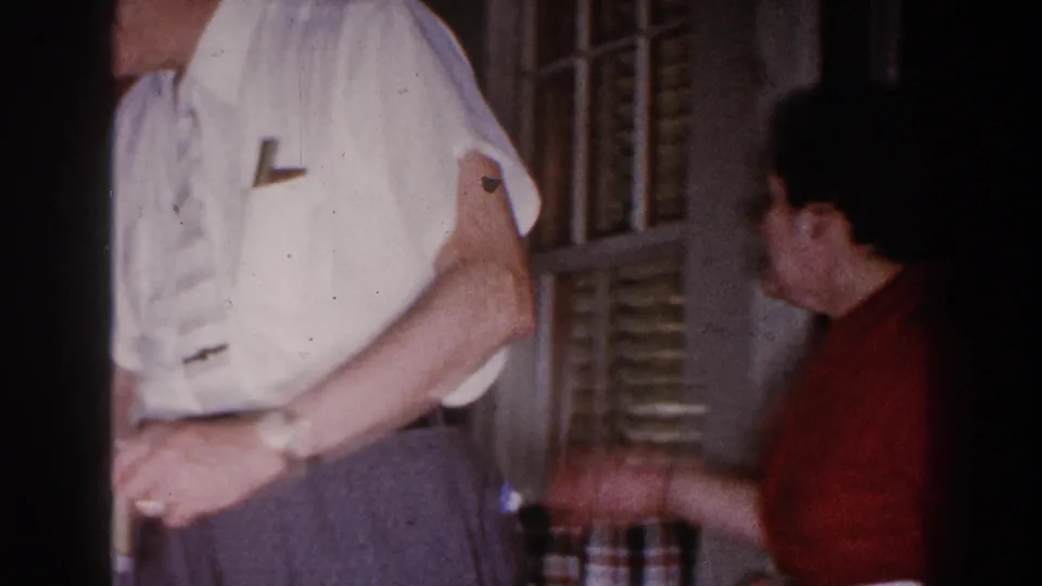 A man in a white shirt and tie stands by louvered windows while a woman in red turns nearby in the home interior.