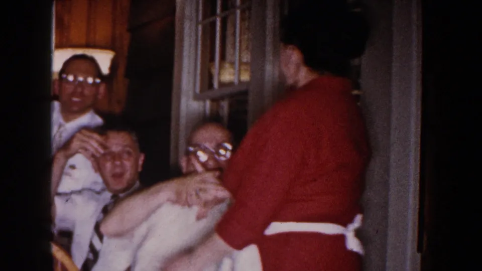 Inside the 1960 Brighton home scene, seated men smile and gesture while the woman in red stands facing them.