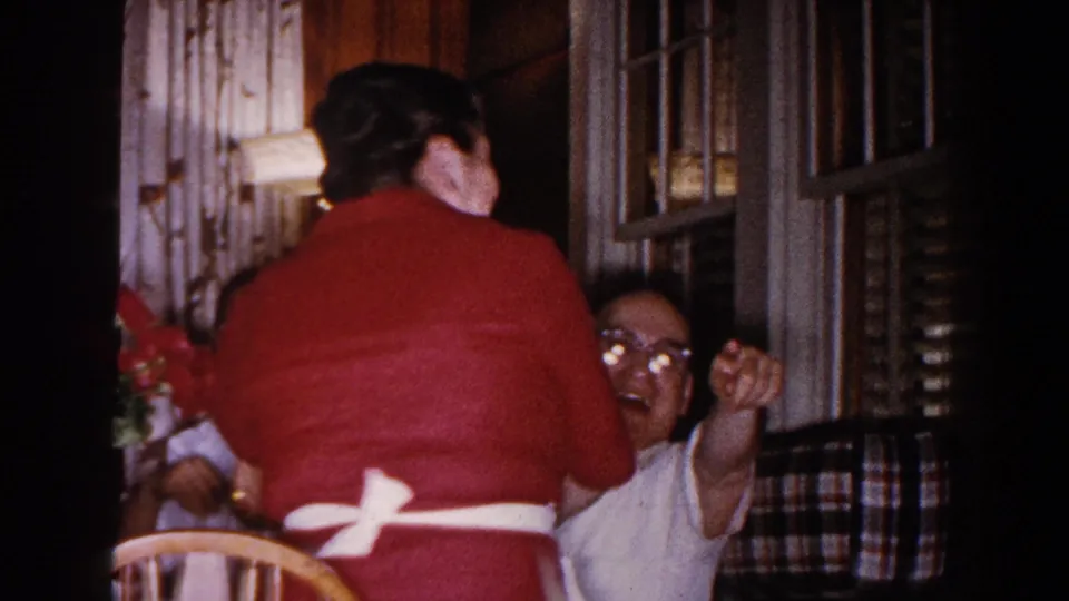 The woman in red stands with her back to the camera as a seated man gestures near louvered windows in the Brighton home interior.