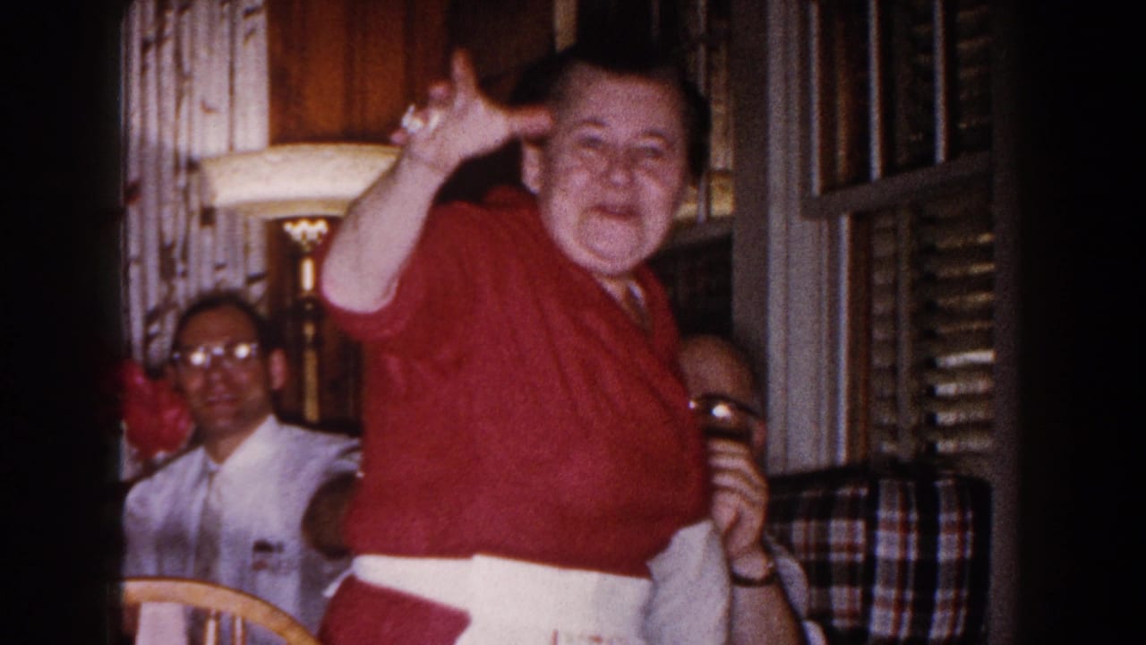 In a 1960 Brighton, Michigan home-movie frame, a woman in a red sweater and white apron waves near seated men in a warmly lit room.