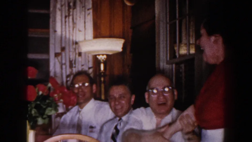 Adults in a Brighton, Michigan interior smile and face the camera, with red roses, a lamp, curtains, and wood paneling visible.
