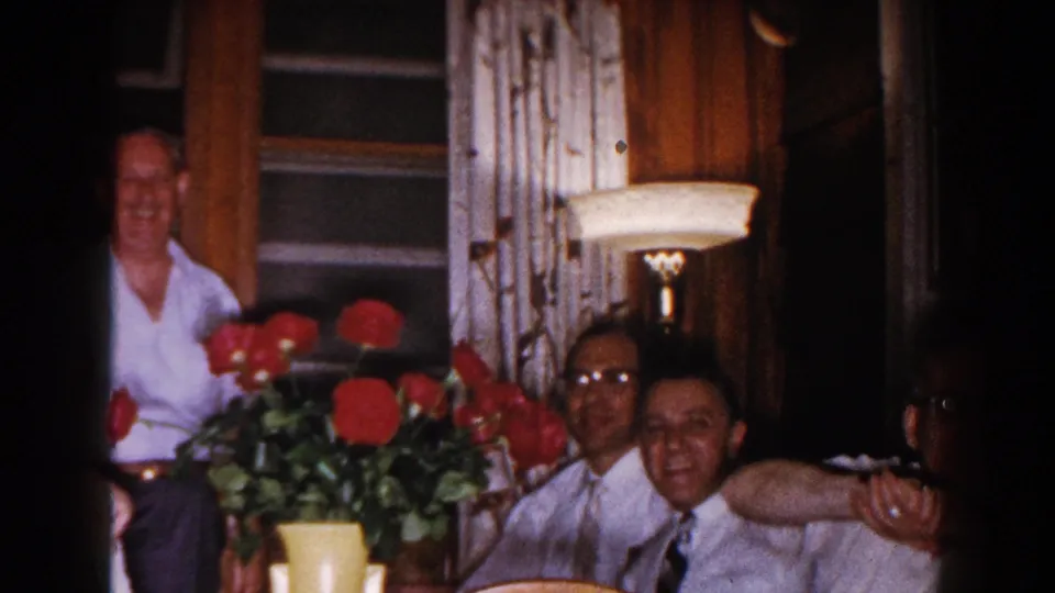 In the home-movie interior, men in light shirts sit and smile near a vase of red roses as another man stands by the window.