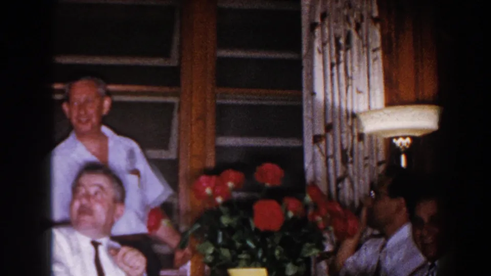 A smiling seated man appears near red roses in a warm Brighton, Michigan interior, with other adults partly visible around him.