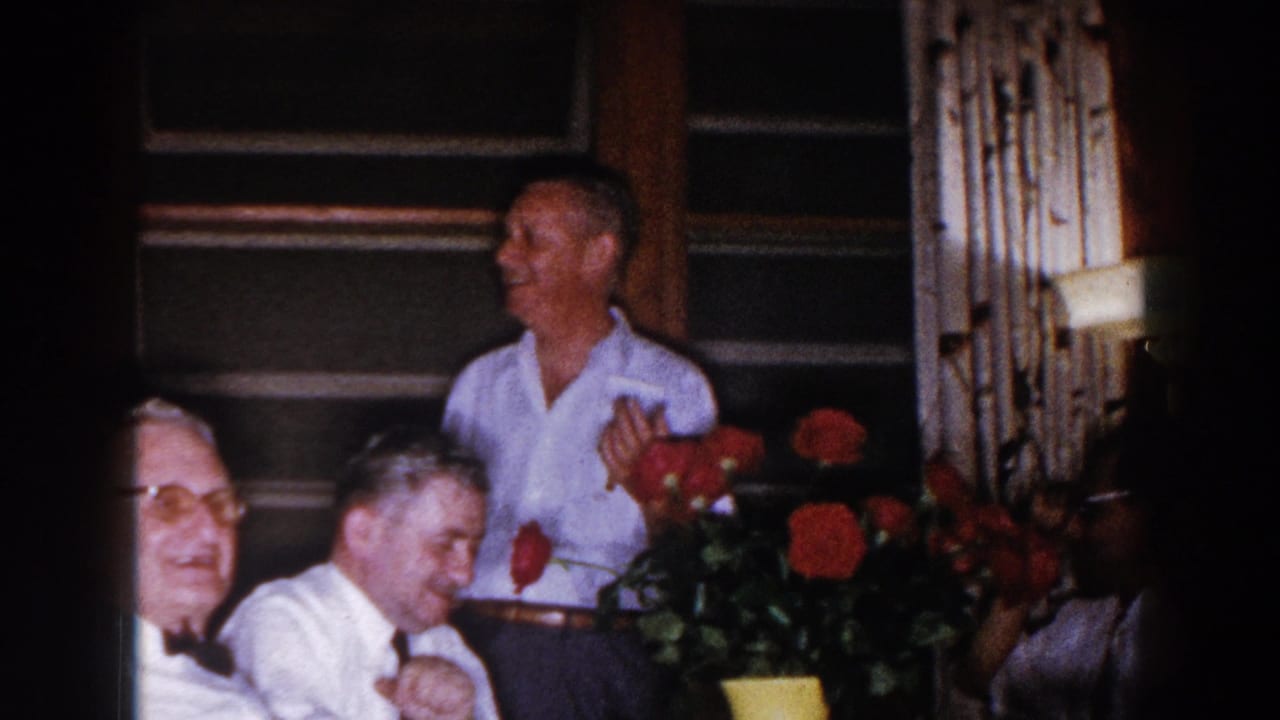 Men in white shirts gather indoors near red roses and wood-paneled windows.