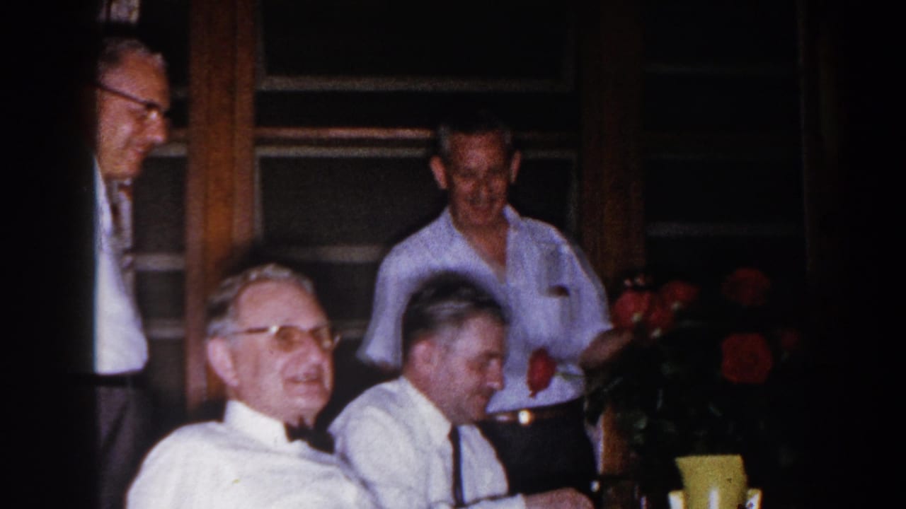 Adult men in white shirts and glasses appear in a Brighton, Michigan indoor gathering with red roses in the foreground.
