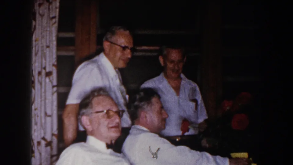 A 1960 home-movie frame shows men in white shirts conversing indoors, with one standing man holding a red rose.