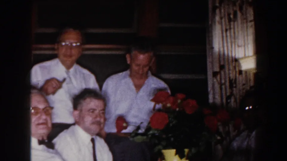 Adult men at a 1960 Brighton, Michigan indoor gathering appear near red roses, with wood paneling and windows behind them.