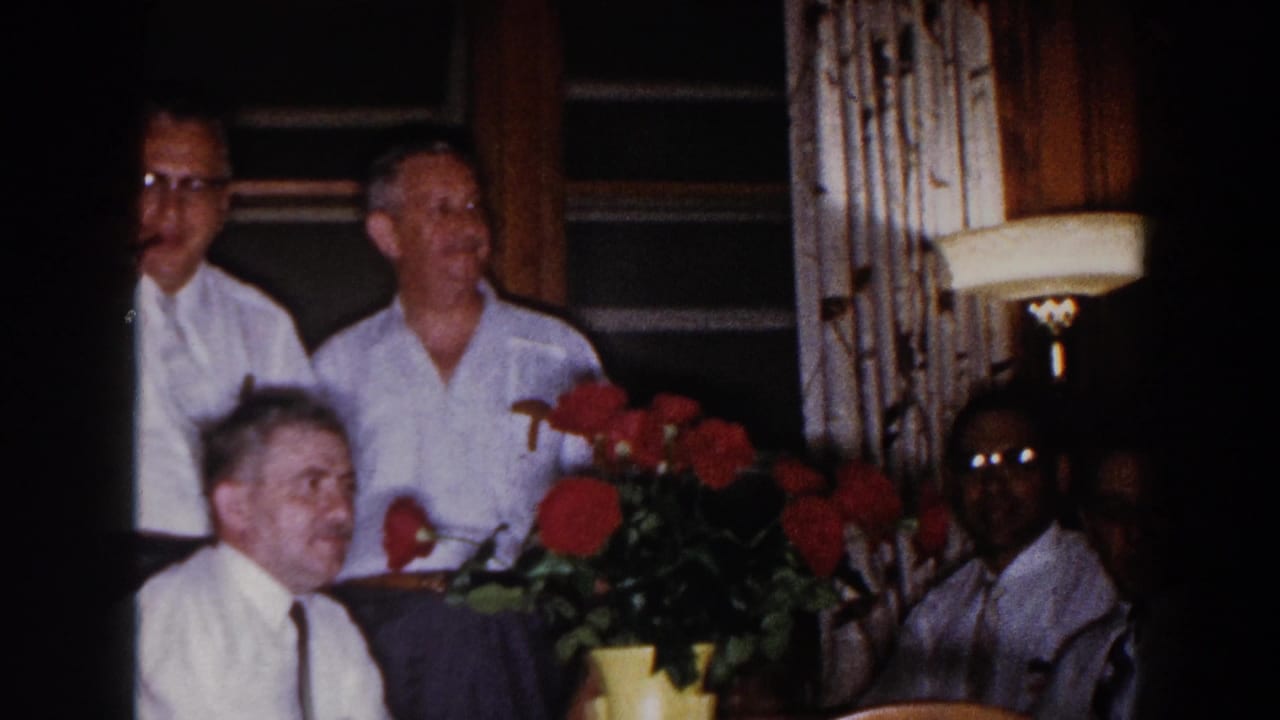1960 Brighton, Michigan home-movie frame of adult men in white shirts gathered around a dining table with red flowers and a lamp nearby.