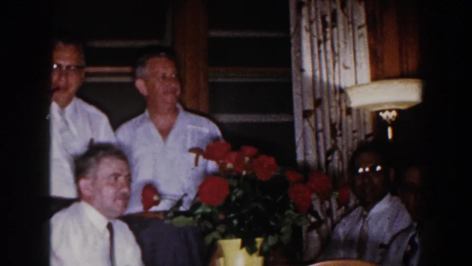 1960 Brighton, Michigan home-movie frame of adult men in white shirts gathered around a dining table with red flowers and a lamp nearby.