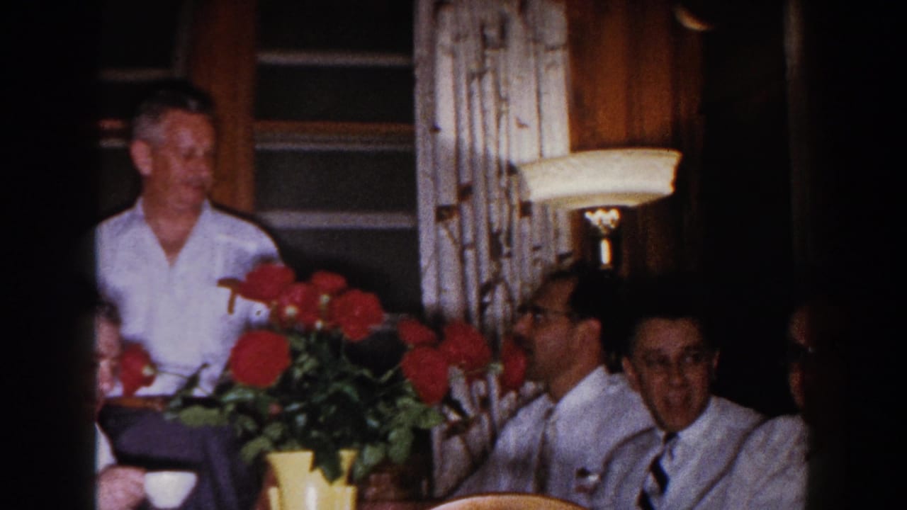 Men at a table look toward the camera beside a red flower arrangement and lit lamp.