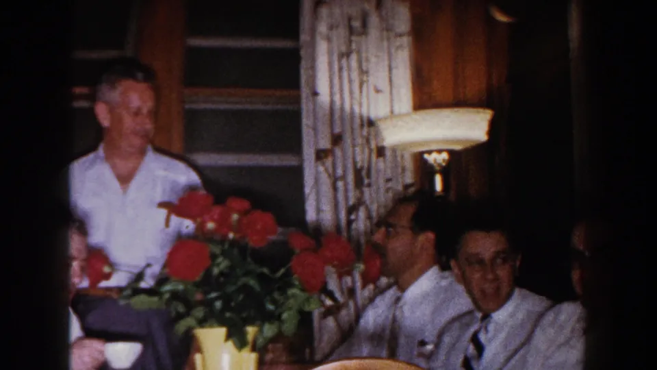 Adult relatives sit and stand near a table in a warm interior, with red flowers, patterned curtains, and a lamp visible.