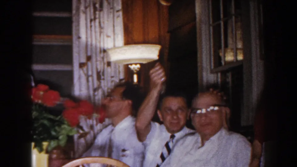 1960 home-movie frame from Brighton showing seated adult men reacting playfully around the dining table in a private interior.