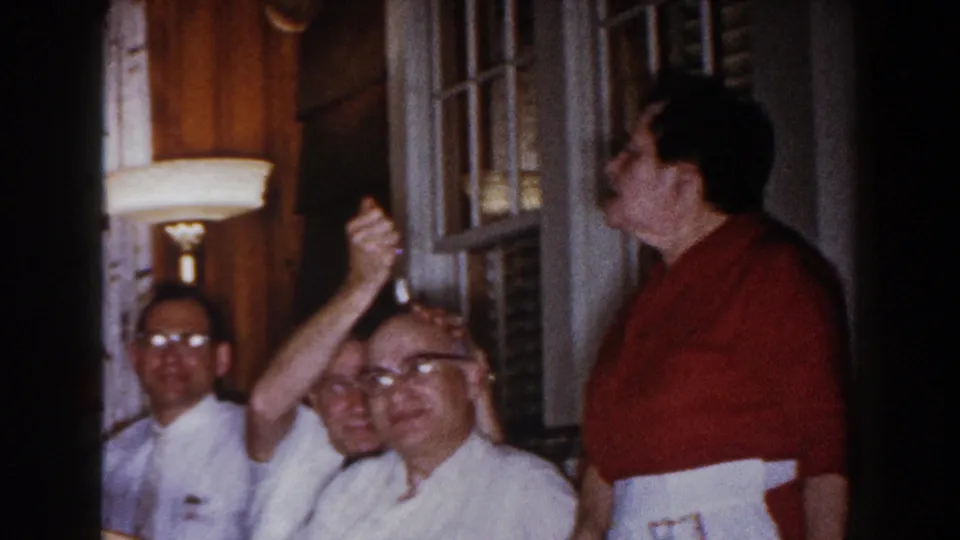 Adult men in a Brighton, Michigan home interior, including a standing man in a red shirt and seated relatives near a lamp and window.