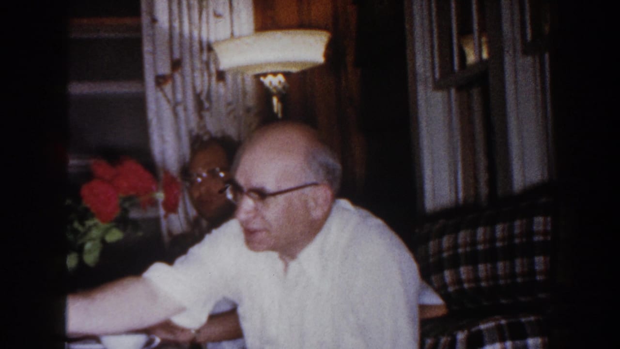 Older man in a white shirt reaches across a table beside red roses.