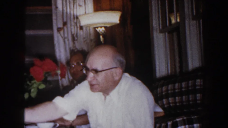An older man in glasses and a white shirt reaches across a dining table during the 1960 Brighton, Michigan family gathering.