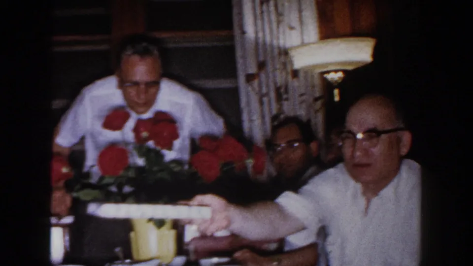 Adults sit close together at a table with red roses in a yellow container, filmed in a softly lit 1960 Brighton, Michigan interior.