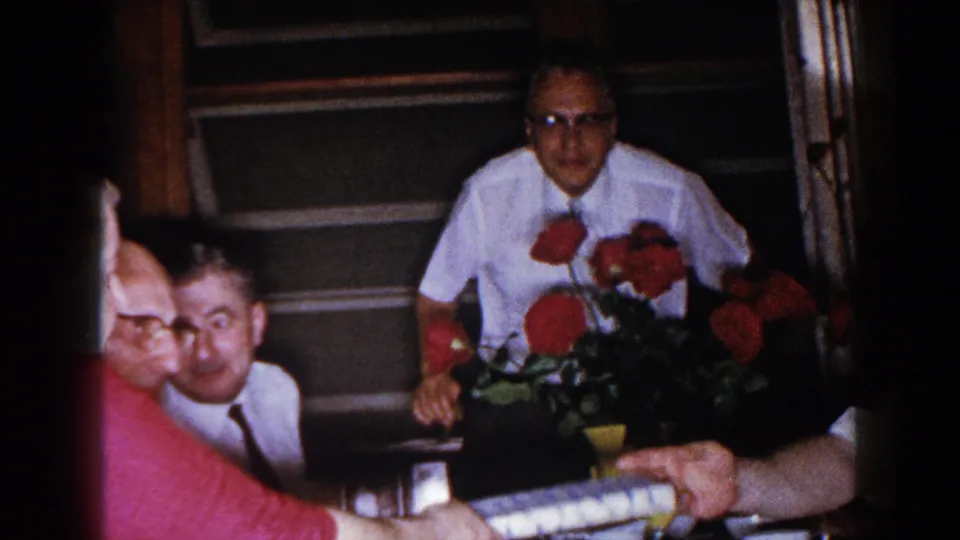A man in a white shirt appears behind red roses at the dining table, with other adults partly visible in the 1960 home-movie frame.