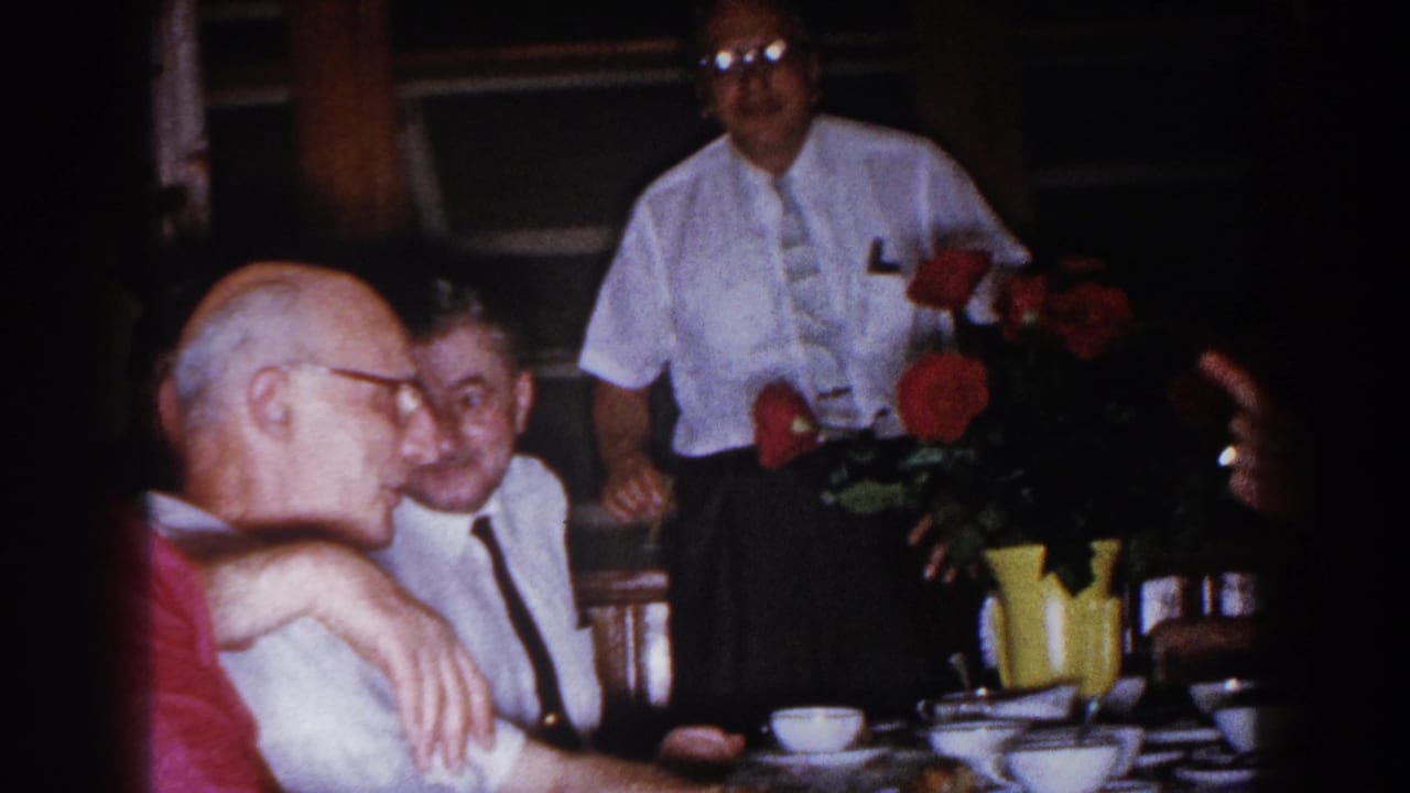 1960 Brighton, Michigan home-movie frame of older men gathered around a set dinner table with red flowers and a yellow pitcher.