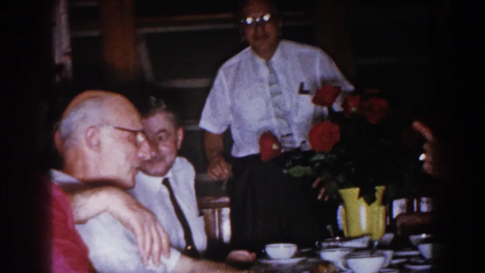 1960 Brighton, Michigan home-movie frame of older men gathered around a set dinner table with red flowers and a yellow pitcher.