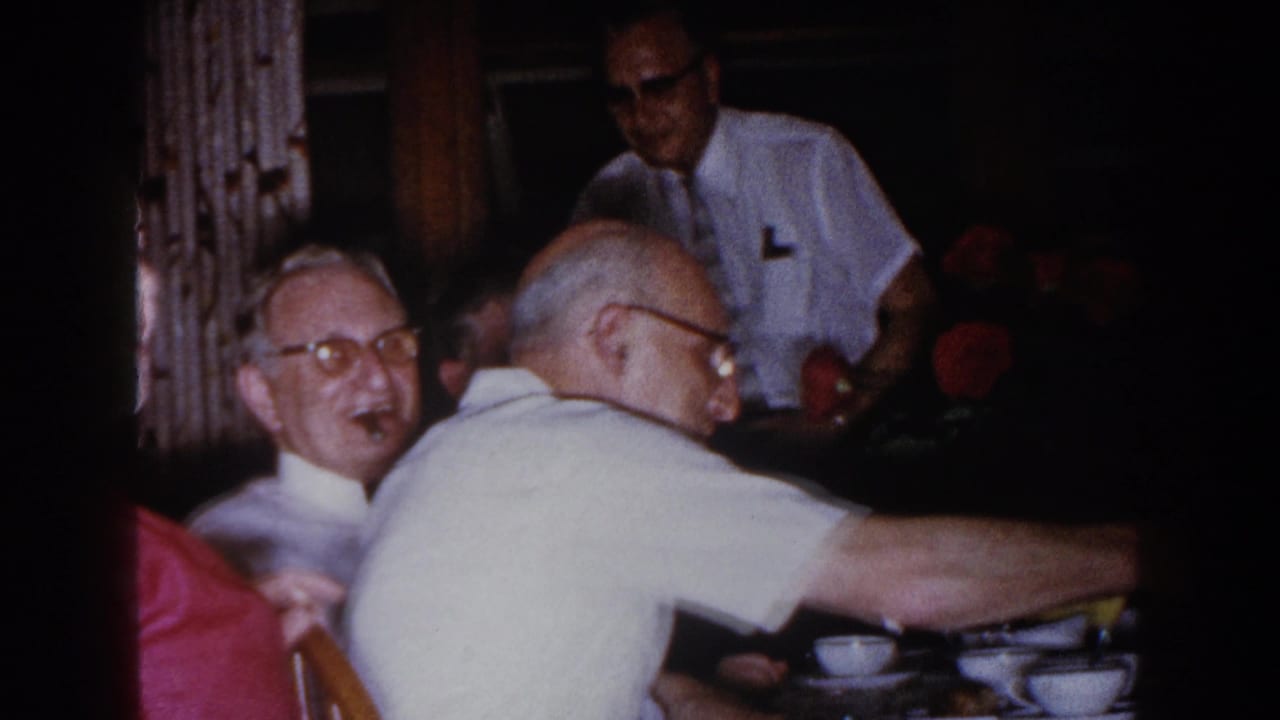 Two older men lean toward each other beside a table with dishes and flowers.