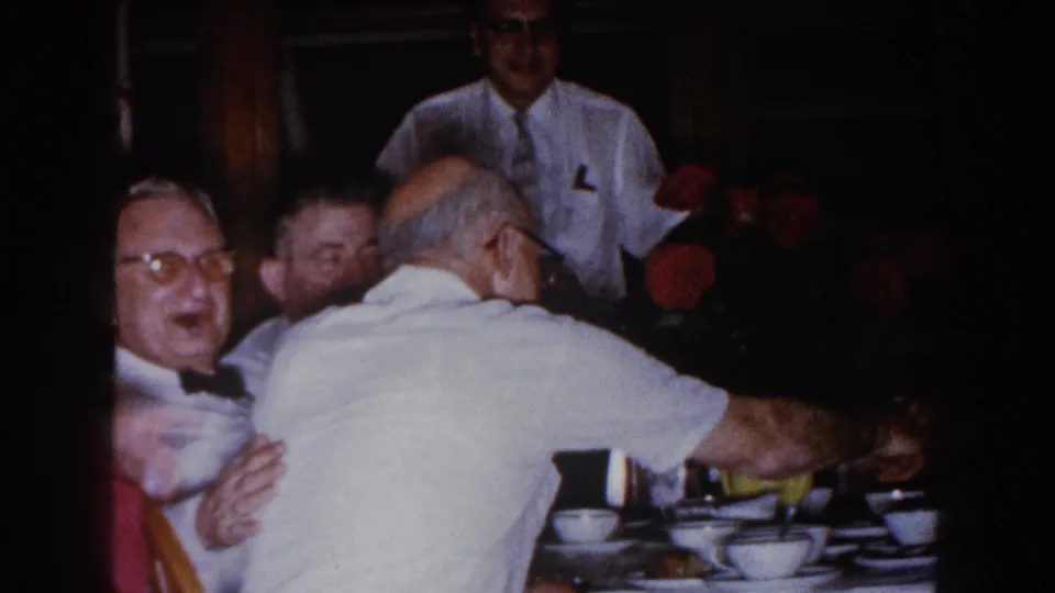1960 Brighton, Michigan home-movie frame of older men seated closely at a crowded dinner table with cups, bowls, and flowers.