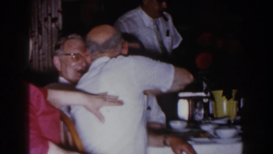 1960 Brighton, Michigan frame showing a friendly embrace among older men seated at a dinner table in a private home.