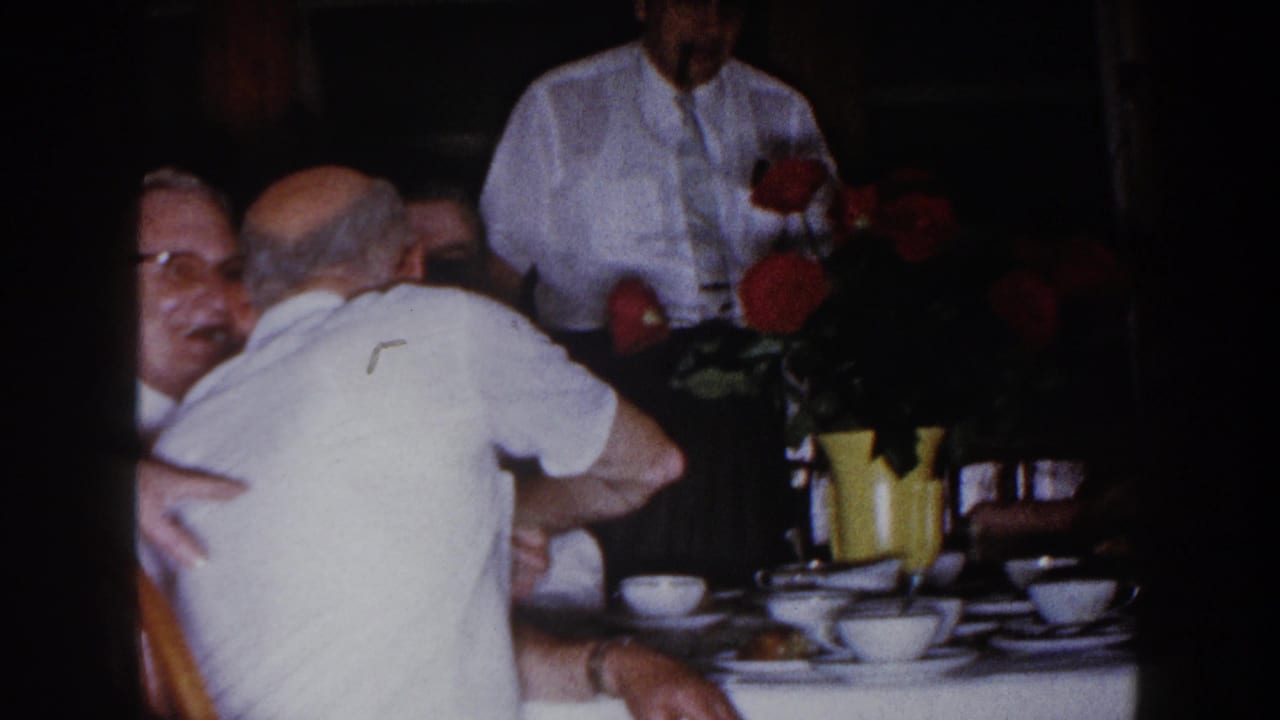 Men seated around a dinner table with cups, a yellow vase, and red flowers in a dim interior.