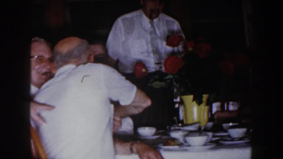 Brighton, Michigan 1960 home-movie frame of adult men at a table with white cups, yellow vase, and red flowers.