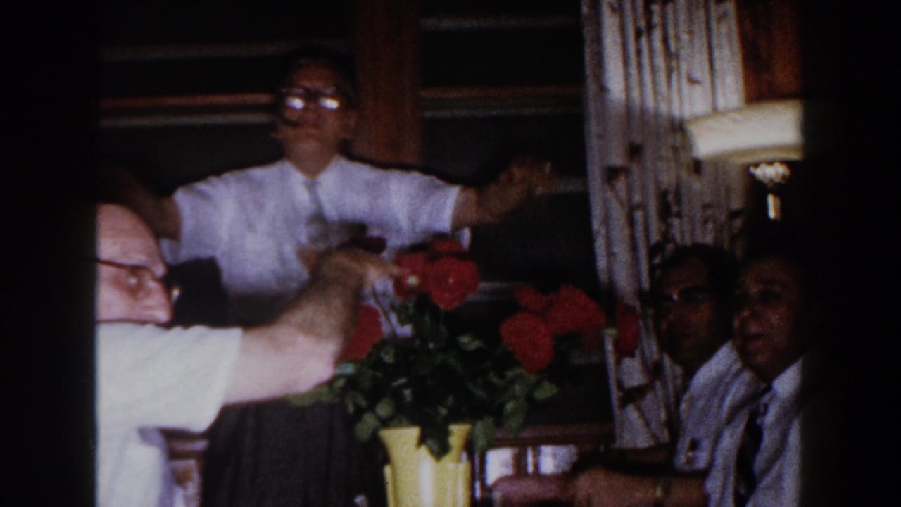 Man in a white shirt reaches across a table near red flowers as other men look on.