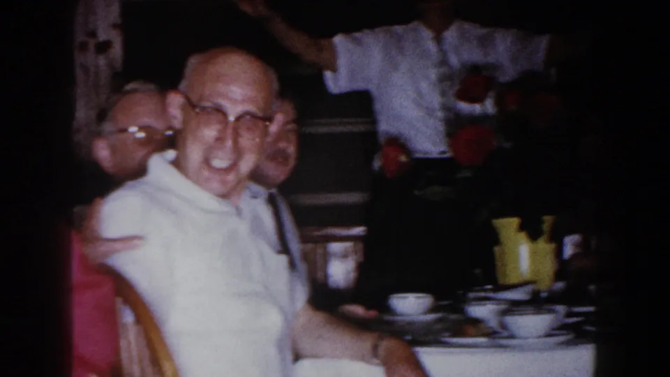 Brighton, Michigan 1960 home-movie frame of an older man in glasses smiling beside a dinner table with cups and flowers.