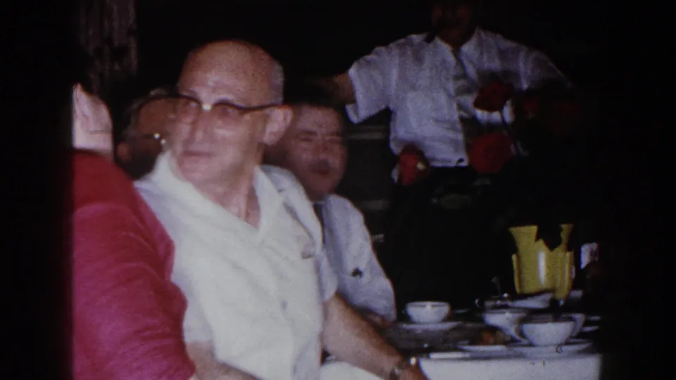 Brighton, Michigan 1960 frame of an older man in glasses turning at a dinner table with cups, flowers, and nearby guests.