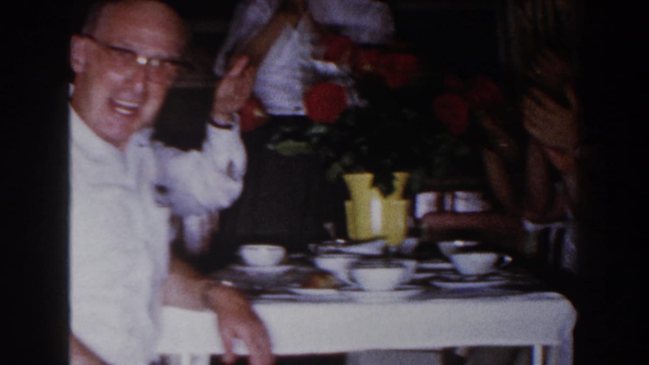 Smiling older man in glasses raises a cloth near a table with cups and flowers.