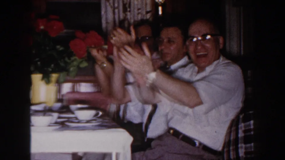 Family members clap at the dinner table, with red flowers, white teacups, and seated adults visible in the 1960 Brighton, Michigan footage.