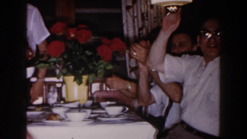At the family dinner in Brighton, Michigan, a man raises his arm as others clap near red flowers and tableware in the 1960 home movie.