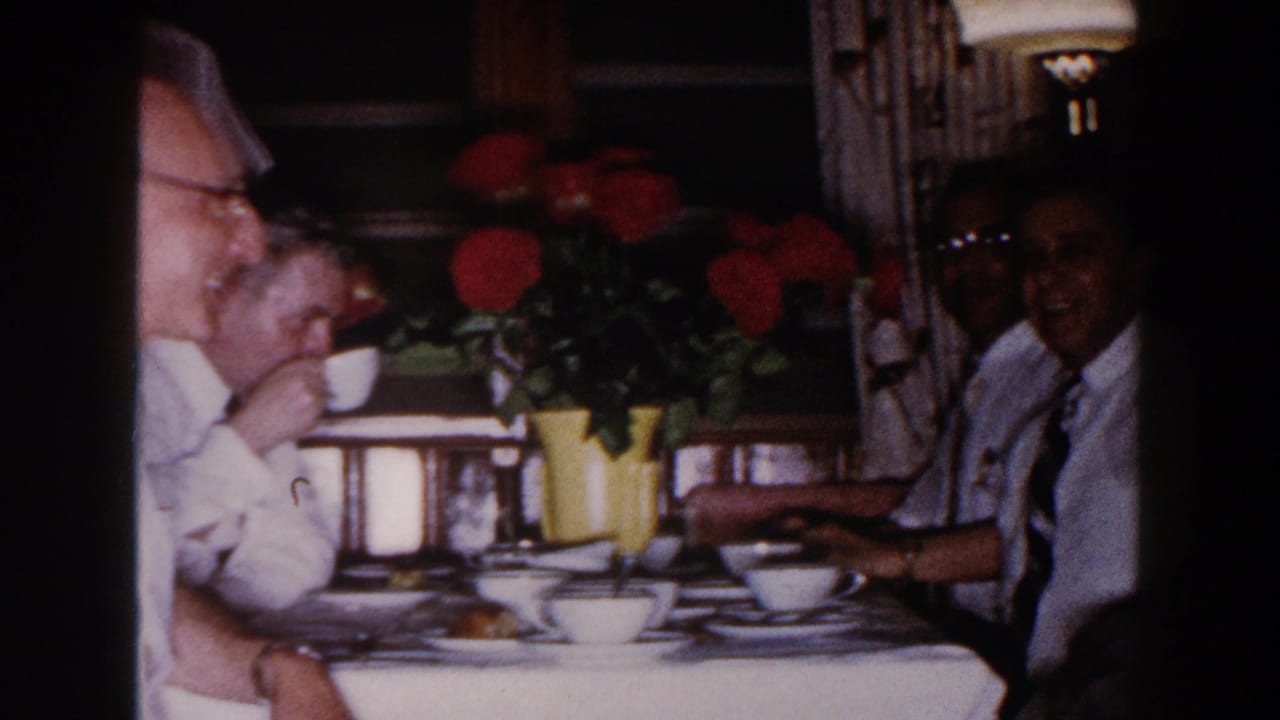 Adults seated at a dining table with cups, plates, pastries, and red flowers in yellow vases.