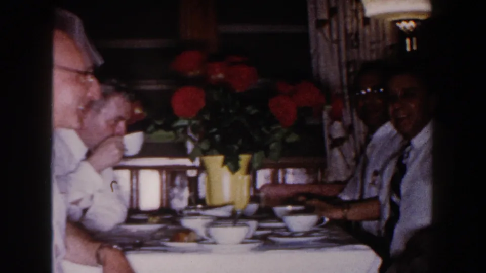 1960 Brighton, Michigan home-movie frame showing adults at a dining table with cups, plates, pastries, and red roses as a centerpiece.
