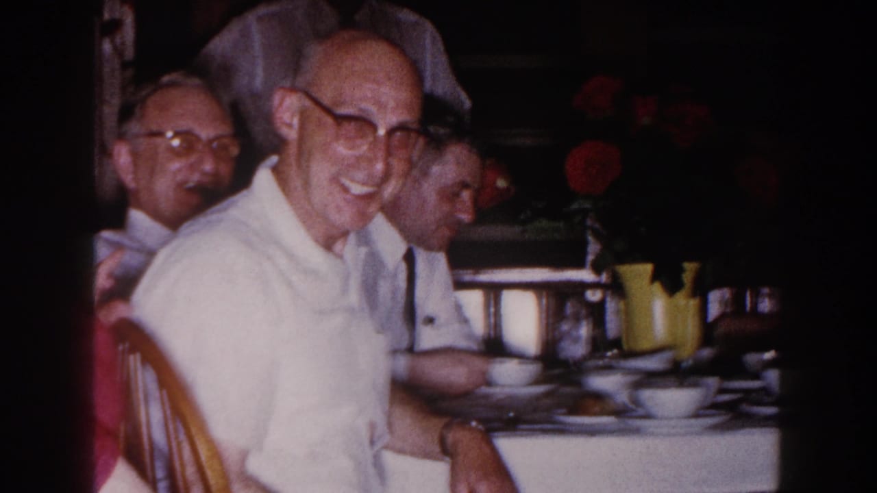 1960 Brighton, Michigan home-movie frame of a smiling man in glasses at a dining table, with other adults and floral decor nearby.