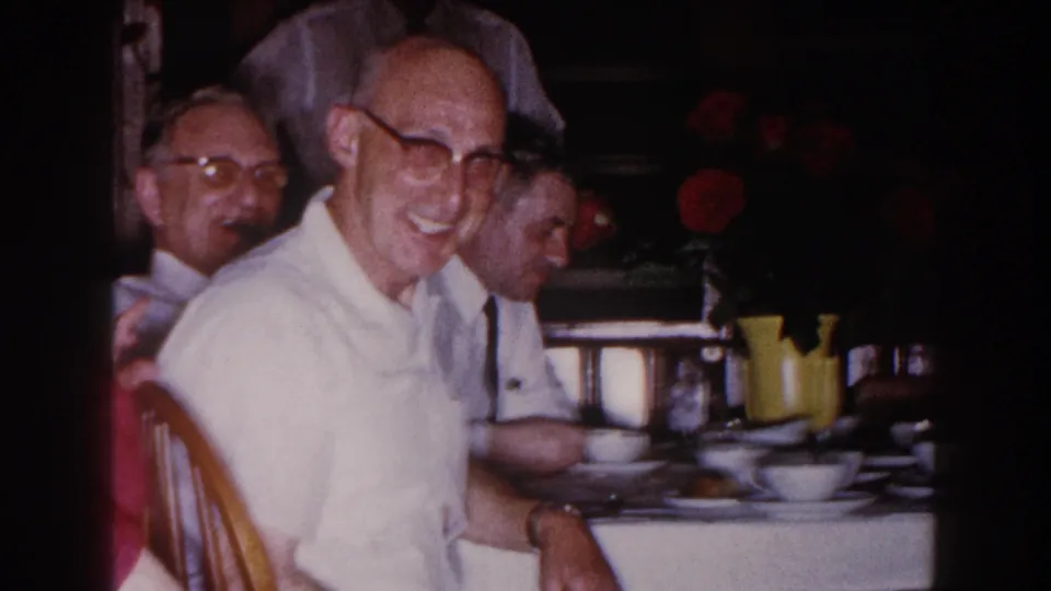 1960 Brighton, Michigan home-movie frame of a smiling man in glasses at a dining table, with other adults and floral decor nearby.