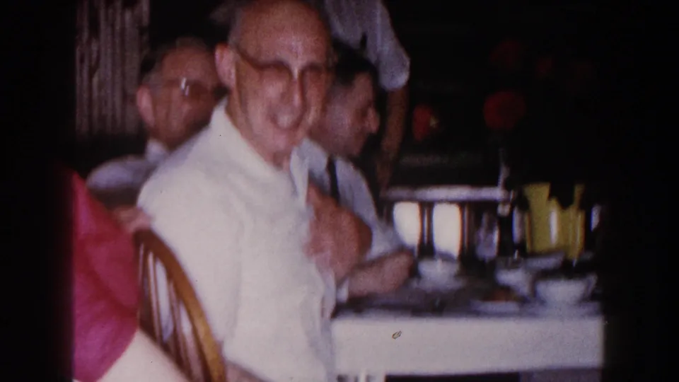 1960 Brighton, Michigan home-movie frame with a softly blurred man in glasses seated at the dining table beside cups and flowers.
