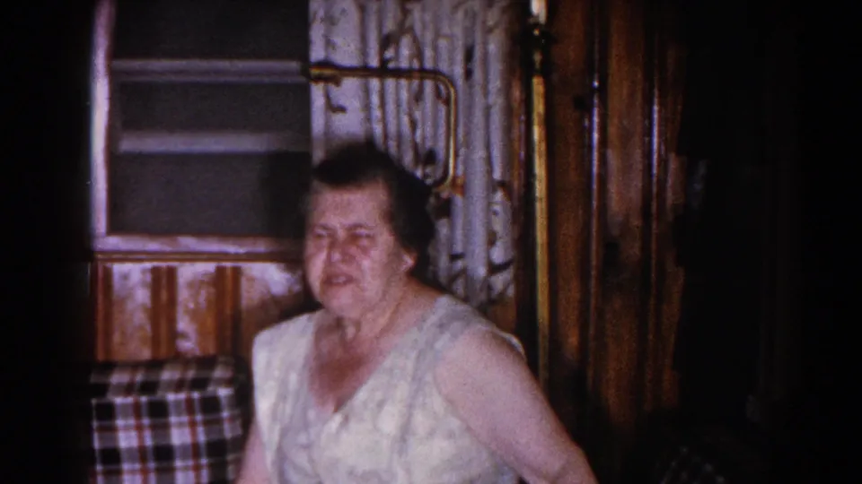 1960 Brighton, Michigan home-movie frame showing a woman in a sleeveless dress seated in a wood-paneled private interior.