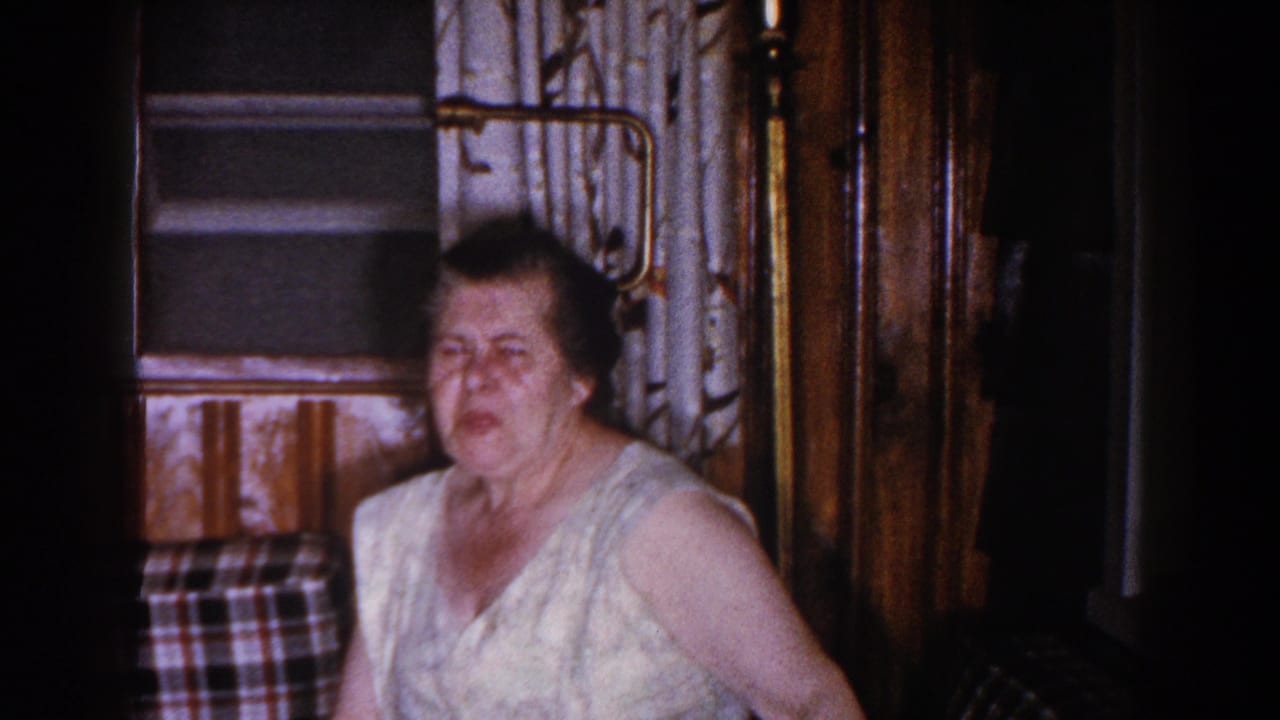 A woman sits indoors in 1960 Brighton, Michigan footage, near patterned curtains, plaid upholstery, and wood paneling.