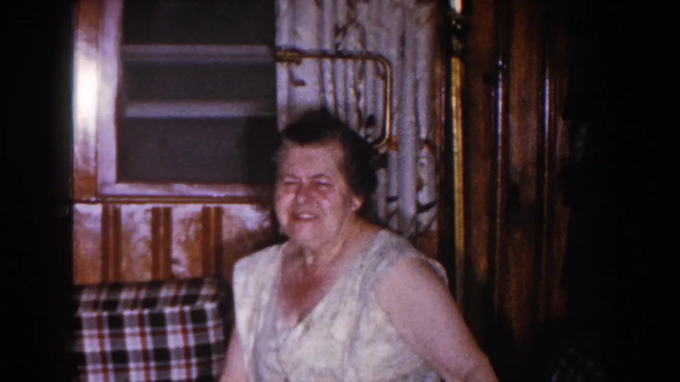 A seated woman smiles or reacts in a dim 1960 home-movie interior with plaid upholstery and patterned curtains.