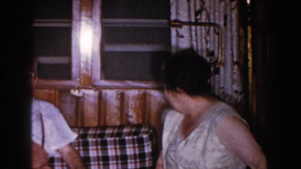The woman turns to the side in the 1960 Brighton home interior, with part of another seated adult visible at the left edge.