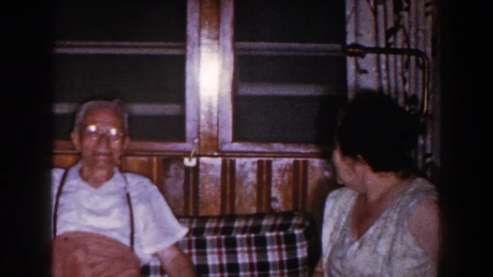 In a 1960 Brighton, Michigan home interior, an elderly man in glasses and suspenders sits on a plaid couch as a woman turns toward him.