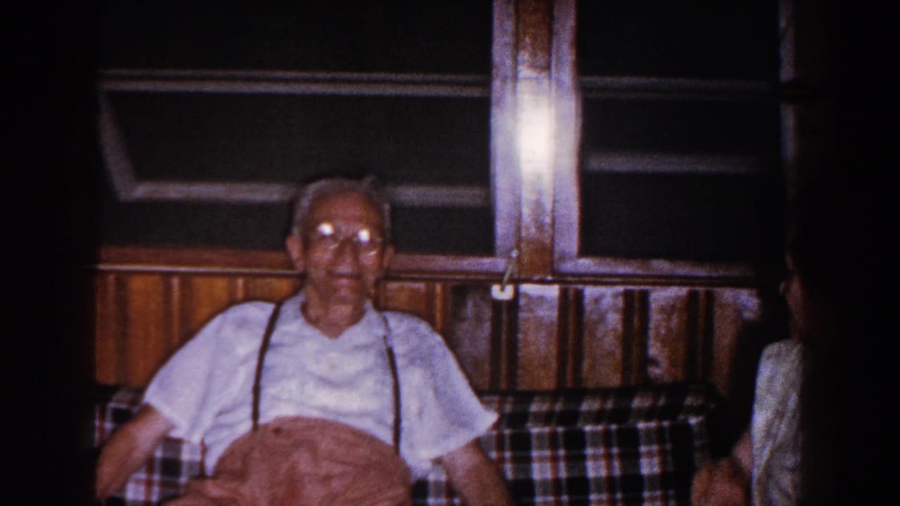 Elderly man sits centered on the couch as a woman is partly visible at the right edge.