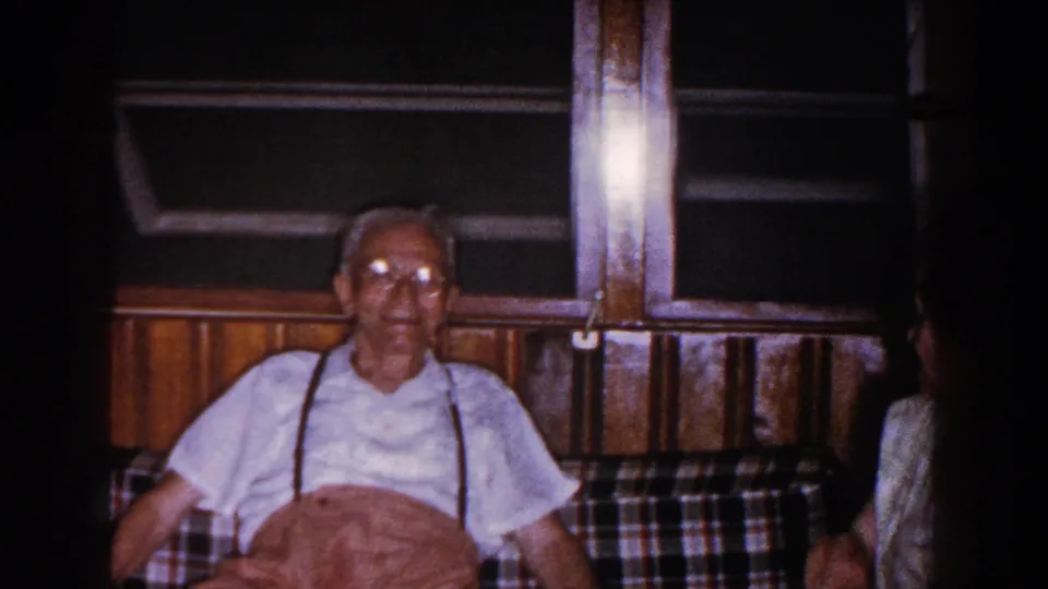 The elderly man remains seated on the plaid couch in the Brighton home interior, with the woman partially cropped at the right side of frame.