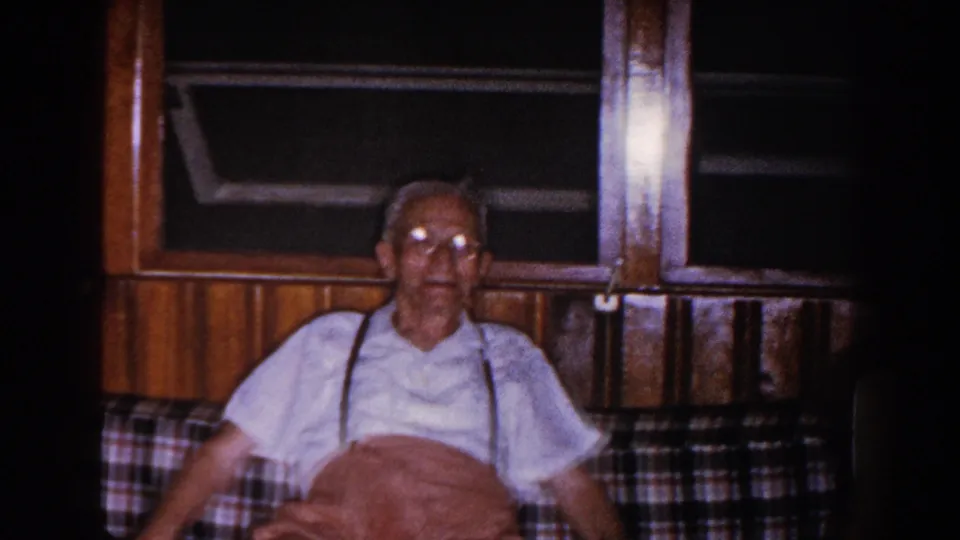 A 1960 home-movie frame shows the elderly man leaning back on the plaid couch, framed by wood paneling and dark windows behind him.