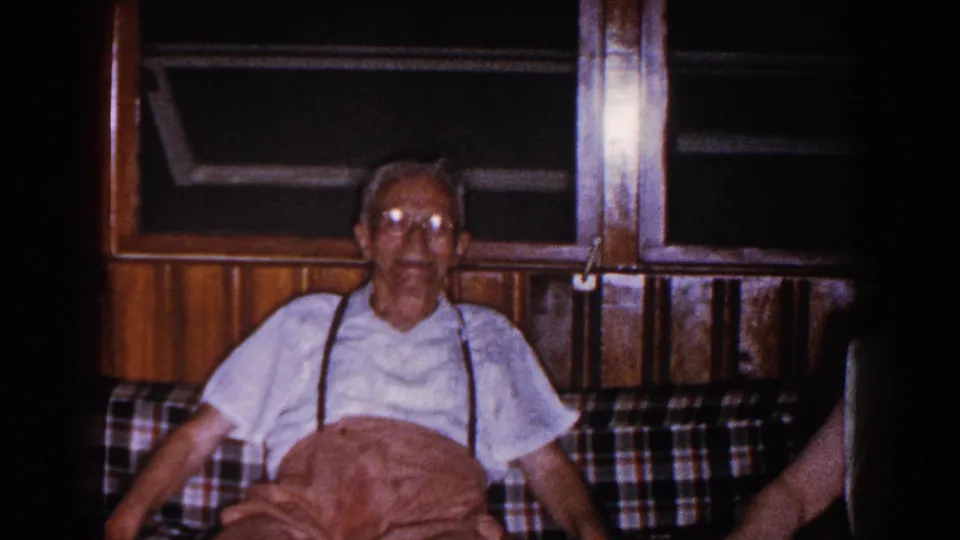 The elderly man sits on the plaid couch as a hand enters from the right, pointing toward him inside the 1960 Brighton home interior.